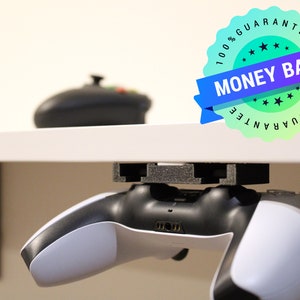 Op de afbeelding: Een witte en zwarte videogamecontroller is gemonteerd onder een wit bureau met een zwarte metalen beugel. De controller wordt op zijn plaats gehouden door een zwarte plastic houder. Een zwarte en groene sticker met de tekst "100% Garantie Geld Terug 100% Garantie" is zichtbaar in de rechterbovenhoek van de afbeelding.