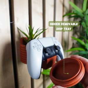 May include: A white video game controller mounted on a wall with a terracotta pot planter. The planter has a removable terracotta drip tray, labeled "HIDDEN REMOVABLE DRIP TRAY".