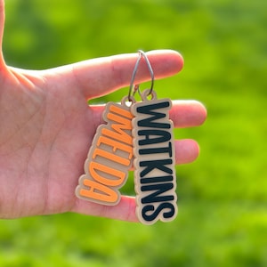 Two personalized name keychains "Imelda" in orange and "Watkins" in black — custom 3D printed bag charm and backpack charm with strong braided cable keyring
