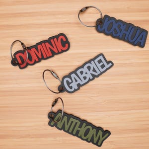 May include: Four keychains with different names on them. The names are Joshua, Dominic, Gabriel, and Anthony. The keychains are made of a soft, flexible material and have a black metal ring.