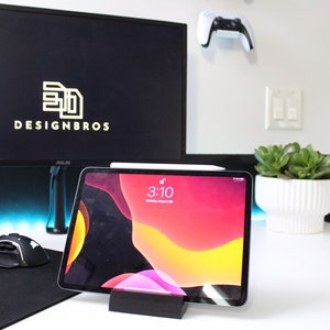 May include: A white desk with a black computer monitor displaying the logo "DESIGNBROS" in gold lettering. A white wireless controller is mounted on the wall behind the monitor. A black tablet with a colorful abstract background is propped up on a black stand. A black mouse and keyboard are on the desk. A white planter with a green succulent plant is on the desk.
