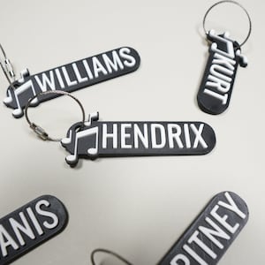 May include: Five black keychains with silver metal rings. Each keychain has a white musical note design and a white name printed on it. The names are "Kurt", "Williams", "Hendrix", "Anis", and "Pitney".