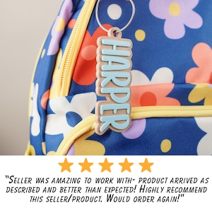 May include: A blue and yellow backpack with a floral pattern. A light blue and brown personalised keychain with the name "HARPER" is attached to the zipper pull.