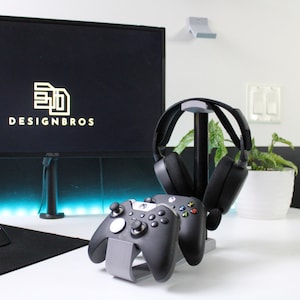 Puede incluir: Un auricular para juegos negro y dos mandos Xbox negros se muestran en un soporte gris. El soporte está sobre un escritorio blanco con una alfombrilla de ratón negra y un ratón negro.