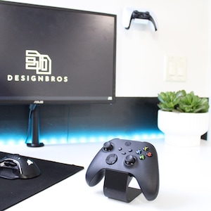 May include: A black Xbox controller on a stand, with a black mouse and keyboard on a black mousepad. The controller is on a white desk with a blue light strip behind it. A white plant pot with a green succulent is on the right side of the image. A white computer monitor with the text "DESIGNBROS" on the screen is in the background.
