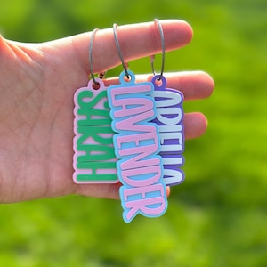 Close-up of three personalized kids keychains in green, pink, and purple — 3D printed custom name tag and diaper bag tag with bold raised lettering outdoors