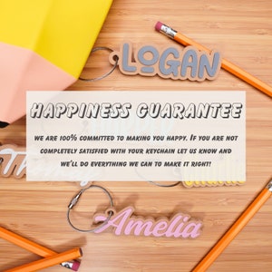 May include: Two personalised wooden keychain tags with the names "Logan" and "Amelia" in grey and pink lettering. The keychain tags are on a wooden surface with pencils. The text "Happiness Guarantee" is on a white rectangle with a peach background. The text below reads "We are 100% committed to making you happy. If you are not completely satisfied with your keychain let us know and we'll do everything we can to make it right!"