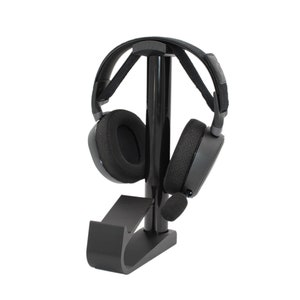 May include: Black headphone stand with a black headset resting on it. The stand has a curved base and a vertical post.