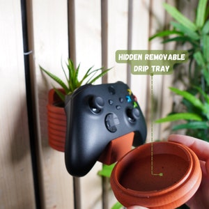 May include: A brown and black plant holder with a removable drip tray. The holder is attached to a wall and has a black video game controller attached to it. The text "HIDDEN REMOVABLE DRIP TRAY" is visible in the image.