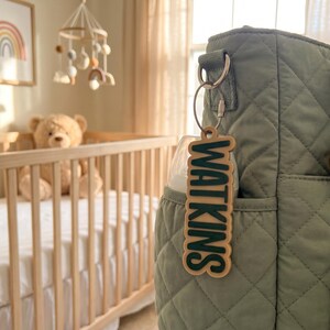 May include: A sage green quilted diaper bag featuring a light-colored wooden keychain with the name WATKINS in dark green raised lettering, attached to a metal ring, with a matching pacifier clipped to the side of the bag.