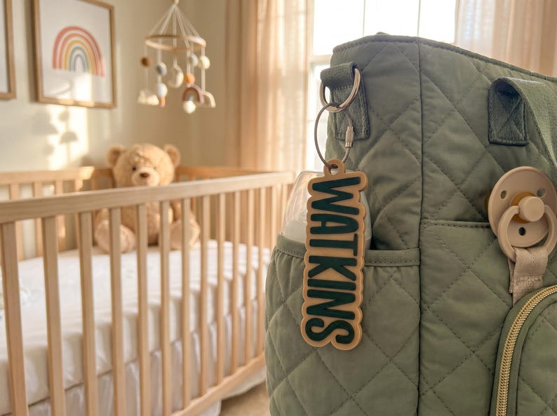 May include: A sage green quilted diaper bag featuring a light-colored wooden keychain with the name WATKINS in dark green raised lettering, attached to a metal ring, with a matching pacifier clipped to the side of the bag.