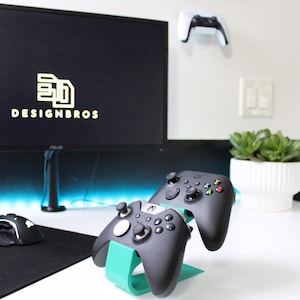 Pode incluir: Dois controladores Xbox pretos são exibidos em um suporte turquesa. Os controladores estão em uma mesa branca com um mousepad preto e um teclado. Um monitor de computador preto com o texto "DESIGNBROS" está no fundo.