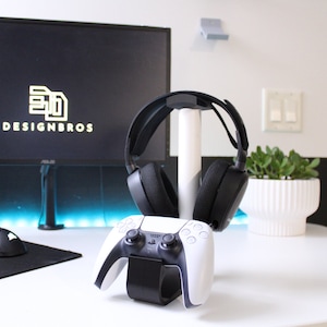 Puede incluir: Un mando de consola de juegos blanco con botones negros y un soporte de auriculares negro con unos auriculares negros en él. Los auriculares están en un soporte blanco. El mando y el soporte de auriculares están en un escritorio blanco.