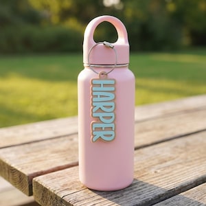 Personalized water bottle tag "Harper" in teal and tan — custom 3D printed name keychain clipped to a pink insulated water bottle outdoors on a picnic table