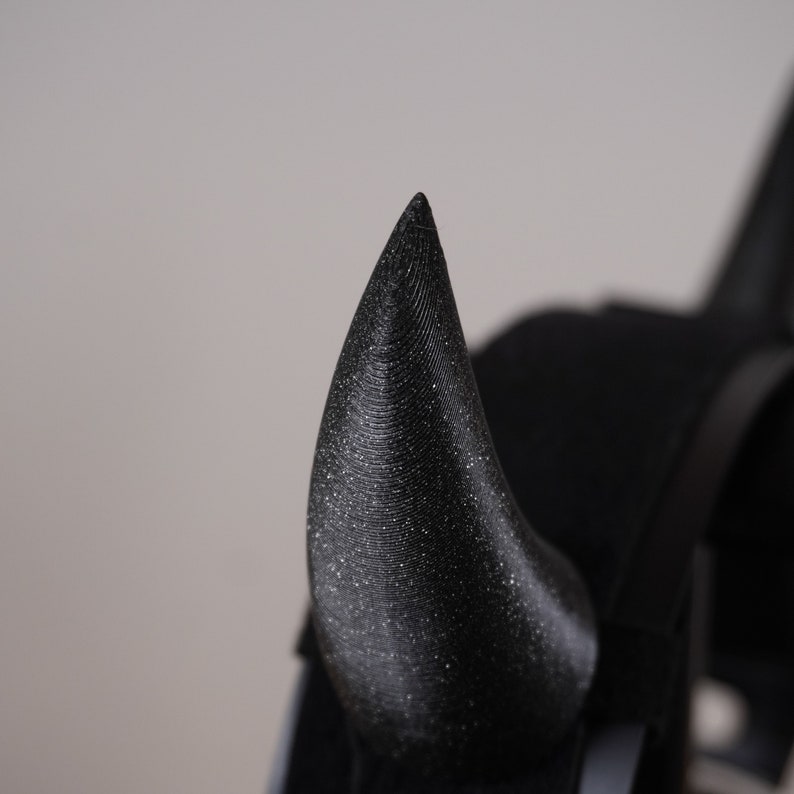 Op de afbeelding: Een close-up van een zwart, glitterend duivelshoorn haarband. De hoorn is puntig en heeft een textuur.