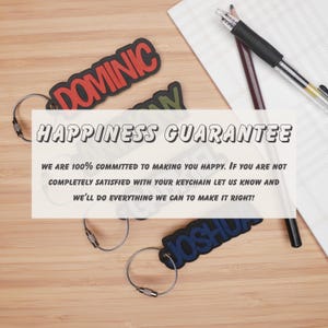 May include: Three personalized keychains with the names Dominic, Josh, and a third name that is partially obscured. The keychains are made of a soft, flexible material and are attached to silver metal key rings. The keychains are displayed on a wooden surface with a white piece of paper and two pencils. The text "HAPPINESS GUARANTEE" is displayed in a white box above the keychains. The text below the box reads "WE ARE 100% COMMITTED TO MAKING YOU HAPPY. IF YOU ARE NOT COMPLETELY SATISFIED WITH YOUR KEYCHAIN LET US KNOW AND WE'LL DO EVERYTHING WE CAN TO MAKE IT RIGHT!"