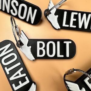 May include: Five black keychains with white text and a white winged shoe charm. The keychains feature the names "BOLT", "LEWIS", "EATON", "INSON", and "[unclear text]".