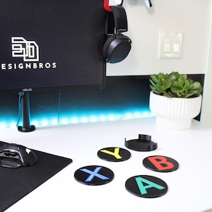 May include: Five round black coasters with colorful letters spelling out "Xbox" on a white desk. The coasters are made of a hard plastic material and have a raised edge. The letters are printed in a bold font and are easy to read. The desk is clean and white, and there is a black mousepad and a computer mouse on the desk.