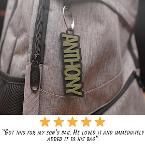 May include: A green and black personalized name tag with the name "ANTHONY" printed on it. The tag is attached to a silver key chain and is hanging from a gray backpack.