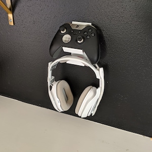 Op de afbeelding: Een witte headset en een zwarte videogamecontroller zijn gemonteerd op een witte wandbeugel.