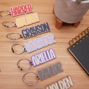 Seven personalized kids keychains in coral, yellow, navy, pink, purple, and grey — custom 3D printed name bag tags with steel cable keyrings on a bamboo desk