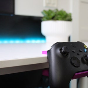 Op de afbeelding: Een zwarte videogamecontroller met een paarse en zwarte standaard. De controller hangt aan de rand van een wit bureau.