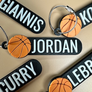 May include: Five black and white keychains with basketball-themed charms. Each keychain features a different name: Giannis, Jordan, Curry, LeBron, and Ko. The basketball charms are orange.