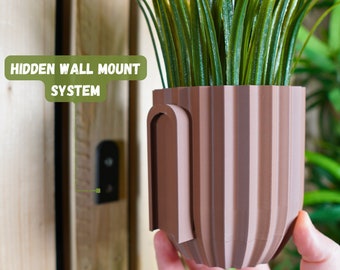 May include: A brown planter with a hidden wall mount system. The planter has a ribbed design and a small opening on the side for mounting. The planter is holding a green plant.