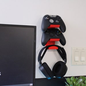 Op de afbeelding: Een rode wandbeugel met twee zwarte Xbox-controllers en een zwarte koptelefoon die eraan hangen. De wandbeugel is bevestigd aan een witte muur.