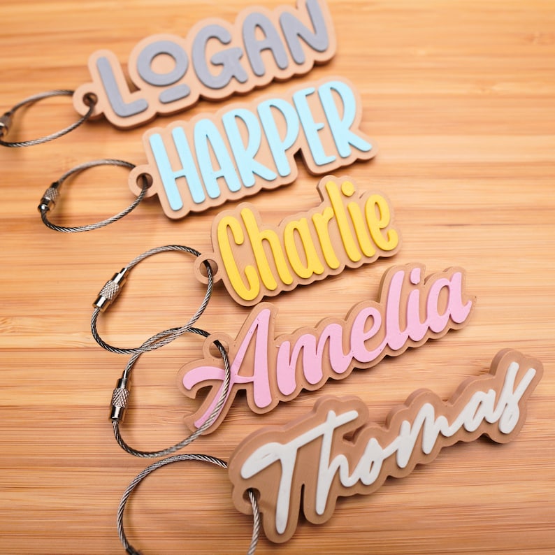 May include: Five personalised keychains with the names Logan, Harper, Charlie, Amelia, and Thomas. Each keychain has a different coloured name on a brown background with a silver metal chain.