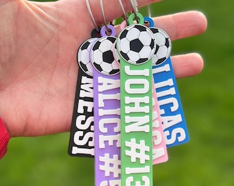 May include: A set of four rectangular plastic bag tags in black, purple, green, and blue, each featuring a soccer ball graphic at the top and white embossed text including the names Messi, Alice #5, John #13, and Lucas, attached to metal loops.