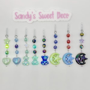 May include: Nine iridescent charm phone charms with different designs, including a heart, teddy bear, star, and crescent moon. The charms are attached to clear plastic strings with iridescent beads.