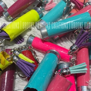 May include: A close-up of a variety of colorful, glittery shotgun shell keychains with tassel accents. The keychains are made from real shotgun shells and are decorated with glitter and colorful paint. The keychains are arranged in a pile on a white fuzzy surface. The text "BROOKE DAWSON'S CREATIONS" is visible in the image.