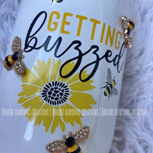 May include: White tumbler with the text "GETTING buzzed" in black and yellow lettering. The tumbler is decorated with a yellow sunflower and several gold bee charms with rhinestones.