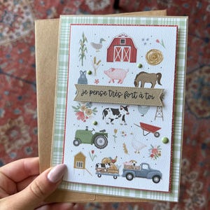 May include: Greeting card with a farm theme. The card features illustrations of a red barn, pig, cow, horse, tractor, and a truck carrying animals. The text "je pense très fort à toi" is on a banner. The card is paired with a brown envelope.