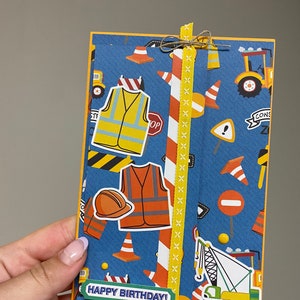 Peut inclure: Carte d'anniversaire artisanale sur le thème de la construction. La carte présente un fond bleu avec des autocollants de construction, notamment des gilets de sécurité, des cônes et des camions. Les mots "Happy Birthday!" sont imprimés sur le devant. Un ruban jaune et de la ficelle ajoutent une touche décorative.