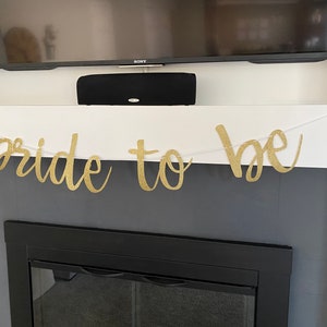 May include: A gold glitter "bride to be" banner hangs above a black fireplace.