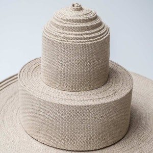 May include: A close-up of a stack of beige cotton webbing rolls. The rolls are stacked in a pyramid shape, with the largest roll at the bottom and the smallest roll at the top.