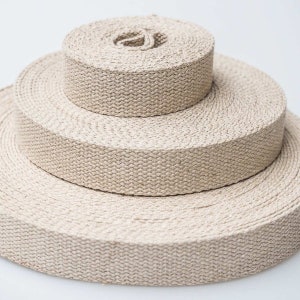 May include: Three rolls of beige woven fabric, each a different size, are stacked on top of each other. The fabric has a textured appearance, and the rolls are tightly wound. The background is plain white.