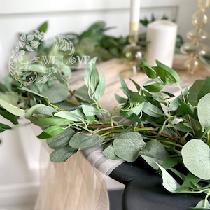 May include: A green eucalyptus garland with leaves and stems, draped over a table with a light brown surface. The garland is ideal for adding a touch of greenery to any space.