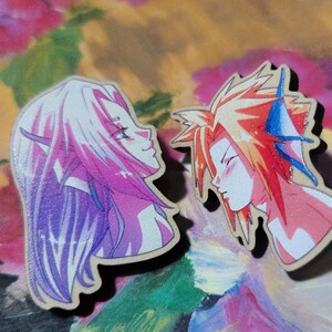 May include: Two wooden pins featuring anime-style characters with detailed facial features and colorful hair. One character has long purple hair and pointed ears, the other has spiky red hair with blue accents. The pins are set against a floral background.