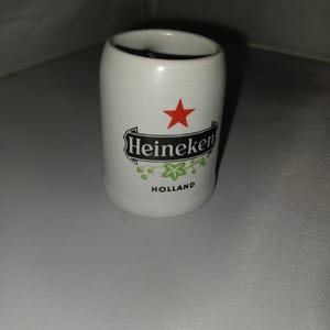 May include: A white ceramic shot glass with the Heineken logo, a red star, and the word "Holland". The shot glass has a cylindrical shape and a smooth surface. The Heineken logo is in black with green accents.
