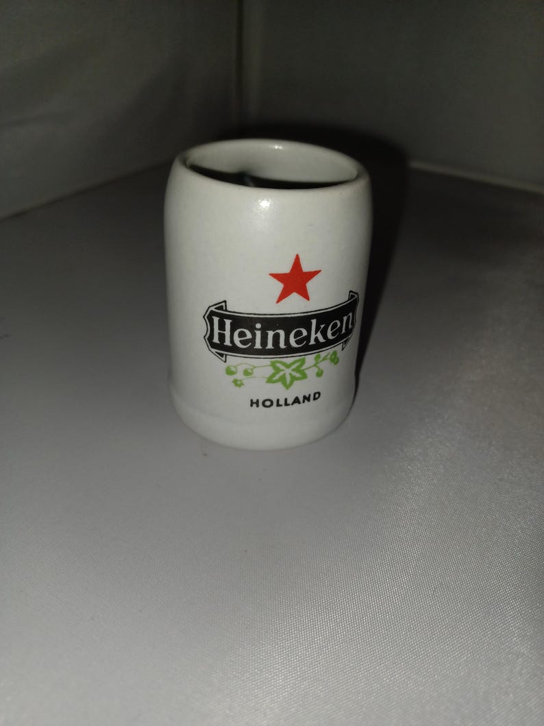 May include: A white ceramic shot glass with the Heineken logo, a red star, and the word "Holland". The shot glass has a cylindrical shape and a smooth surface. The Heineken logo is in black with green accents.