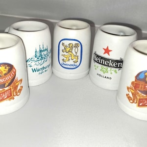 May include: A collection of white ceramic shot glasses. Each glass features a different logo, including a globe with "Schlitz", a lion, a castle, and the Heineken logo with a red star. The glasses are cylindrical with a slightly flared top.