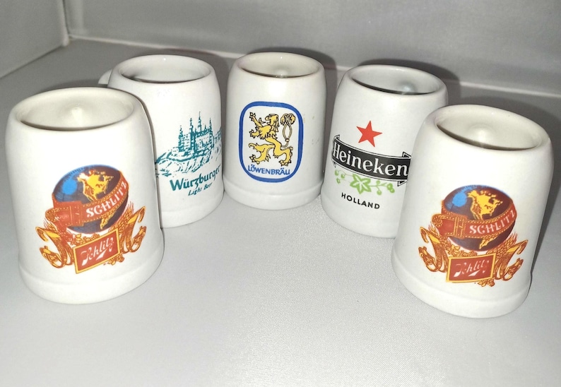 May include: A collection of white ceramic shot glasses. Each glass features a different logo, including a globe with "Schlitz", a lion, a castle, and the Heineken logo with a red star. The glasses are cylindrical with a slightly flared top.