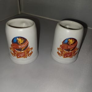 May include: Two white ceramic shot glasses with a curved shape. Each glass features a colorful emblem with a globe, wheat, and the word "Schlitz" in a decorative font. The glasses are likely vintage collectibles.