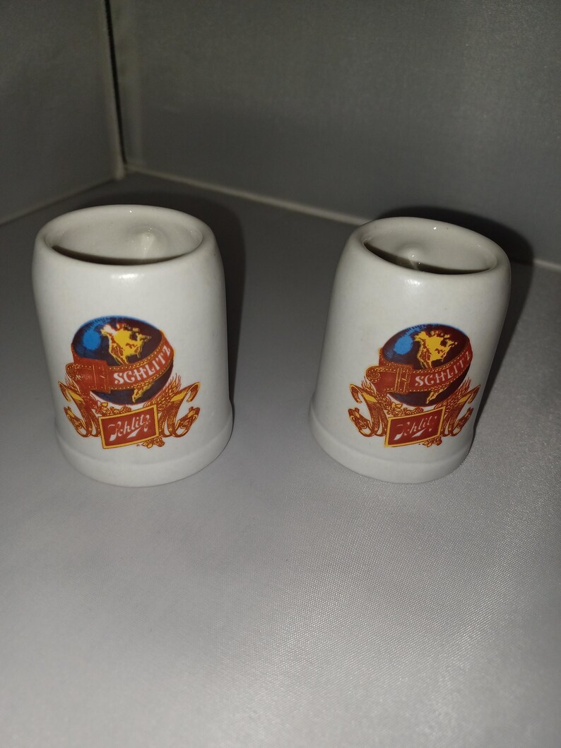 May include: Two white ceramic shot glasses with a curved shape. Each glass features a colorful emblem with a globe, wheat, and the word "Schlitz" in a decorative font. The glasses are likely vintage collectibles.
