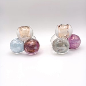 May include: Two decorative glass orb candle holders. Each holder has a clear, round top with a colored orb base. The bases are in shades of blue, purple, and gray. The candle holders are on a white background.