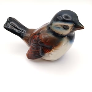 May include: A ceramic bird figurine with a glossy finish. The bird features a dark blue head, brown and white body, and black tail. The figurine is a decorative item.