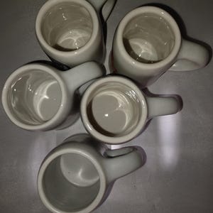 May include: Five small, off-white ceramic mugs with handles are arranged in a cluster. The mugs have a simple, cylindrical shape with a rounded handle. The interior of each mug is visible, and they appear clean. The mugs are set against a neutral background.